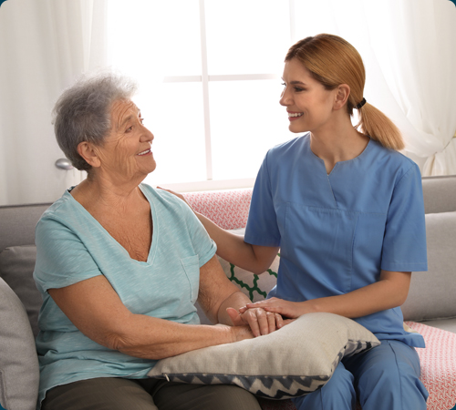 A James River Home Health & Hospice nurse in blue scrubs smiling and holding the hand of an elderly patient on a sofa in Lynchburg, VA.