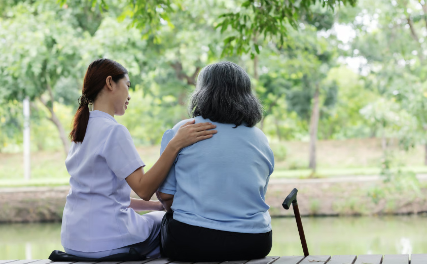 A young female nurse in a light purple uniform sits beside an elderly woman, placing a supportive hand on her shoulder as they look out over a peaceful park pond.