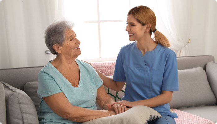 A James River Home Health & Hospice nurse in blue scrubs smiling and holding the hand of an elderly patient on a sofa in Lynchburg, VA.