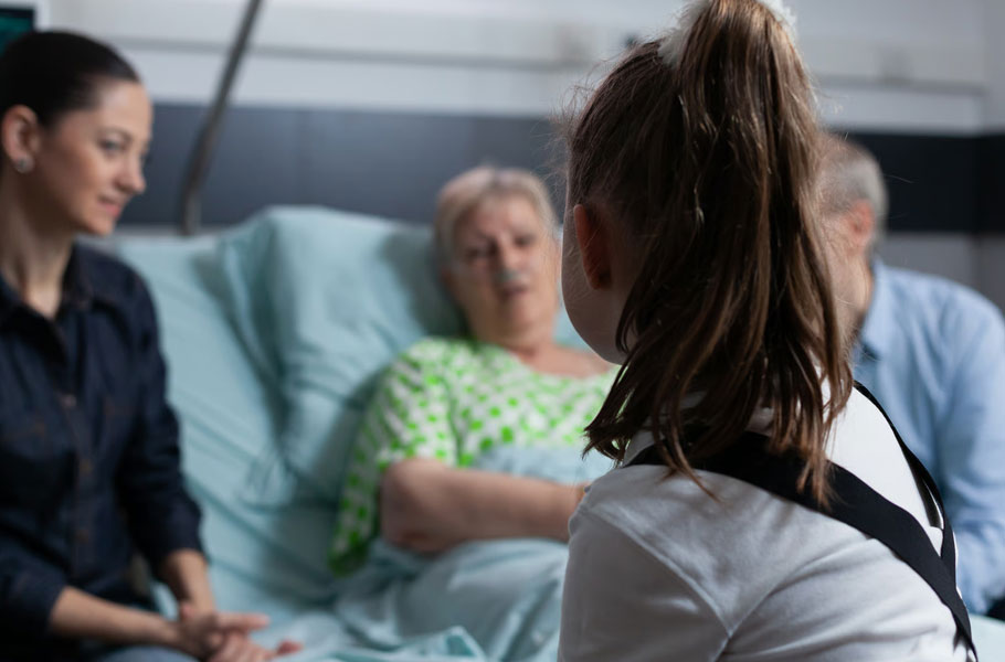 A young girl viewed from behind visits her elderly grandmother resting in a hospital bed with a nasal cannula, while a supportive woman sits bedside, illustrating a family recognizing the signs that it may be time for hospice care in Virginia with James River Home Health & Hospice.