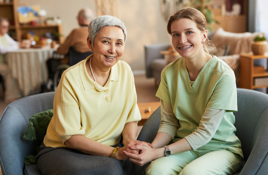 A smiling hospice nurse in green scrubs holding hands with a happy elderly woman in a yellow shirt.