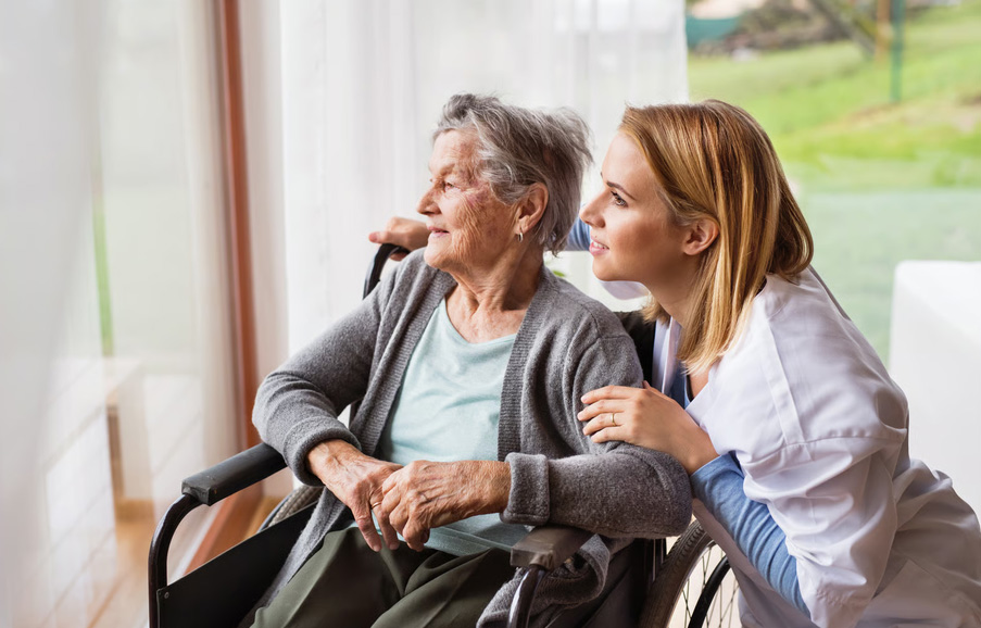 A hospice nurse gently placing a hand on an elderly woman's shoulder as they look out a sunny window together at home.