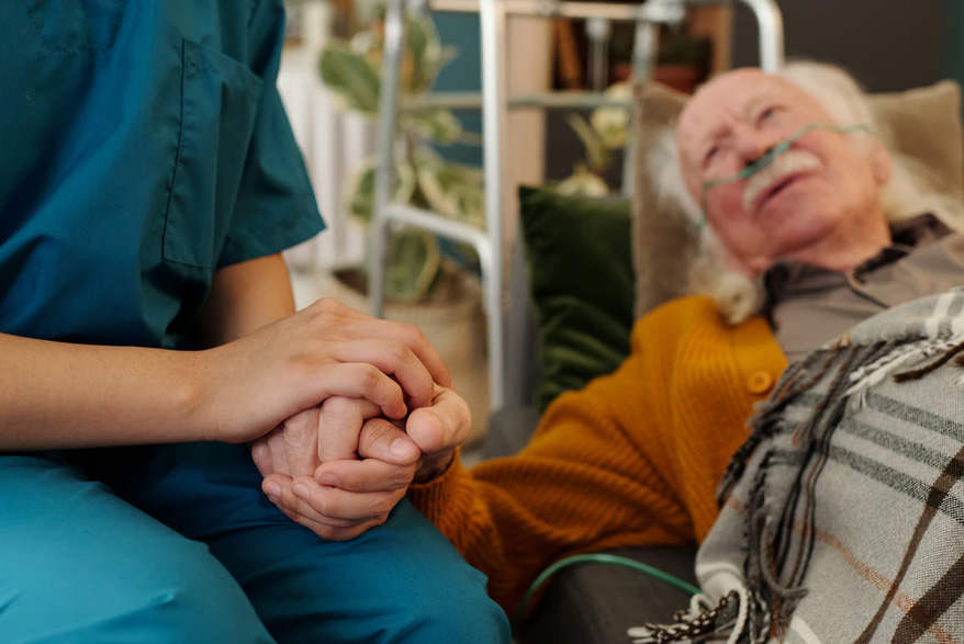 A healthcare worker in teal scrubs holding hands with an elderly male hospice patient resting in bed with nasal cannula.
