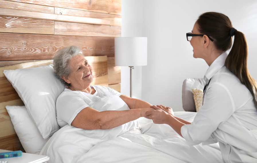 A smiling doctor with glasses holding hands with an elderly woman resting comfortably in bed.