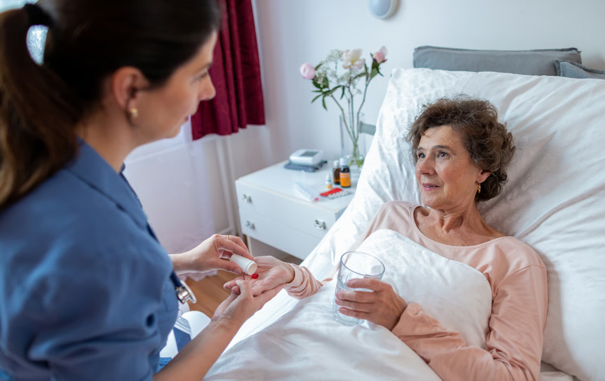 A female nurse in blue scrubs handing medication and a glass of water to a senior woman resting in bed.